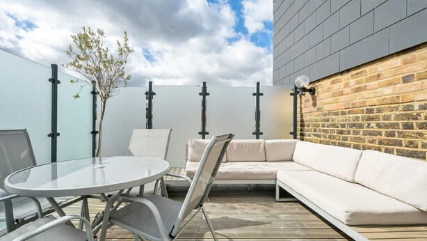 Een terras buiten met een ronde tafel, twee stoelen, een bank en een kleine boom.