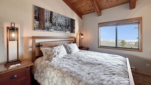 Une chambre à coucher avec un grand lit, une tête de lit en bois, deux tables de chevet avec des lampes, un tableau représentant une forêt et une fenêtre donnant sur un beau paysage.