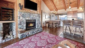 Un salon chaleureux avec une cheminée en pierre, des poutres apparentes en bois au plafond, un téléviseur fixé au-dessus de la cheminée, et un coin repas avec une table et des chaises.