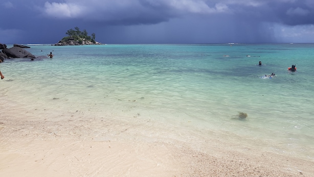 Uma praia com águas turquesa cristalinas, um afloramento rochoso e uma ilha ao longe.