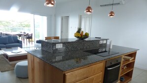 Cozinha moderna com bancadas de granito, armários de madeira e forno embutido. Há uma área de jantar com um sofá azul e uma mesa de centro.