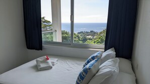 Uma cama com lençóis brancos, um travesseiro azul e branco, e uma toalha dobrada com uma flor vermelha.