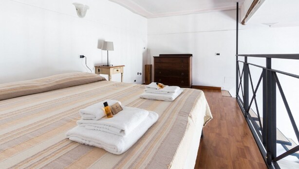 Habitación de hotel con una cama grande, un aparador de madera, una mesita de noche con lámpara y un balcón con baranda de metal.