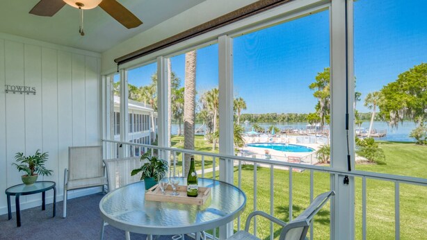 A room with a glass table, chairs, a ceiling fan, and a view of a pool and greenery.