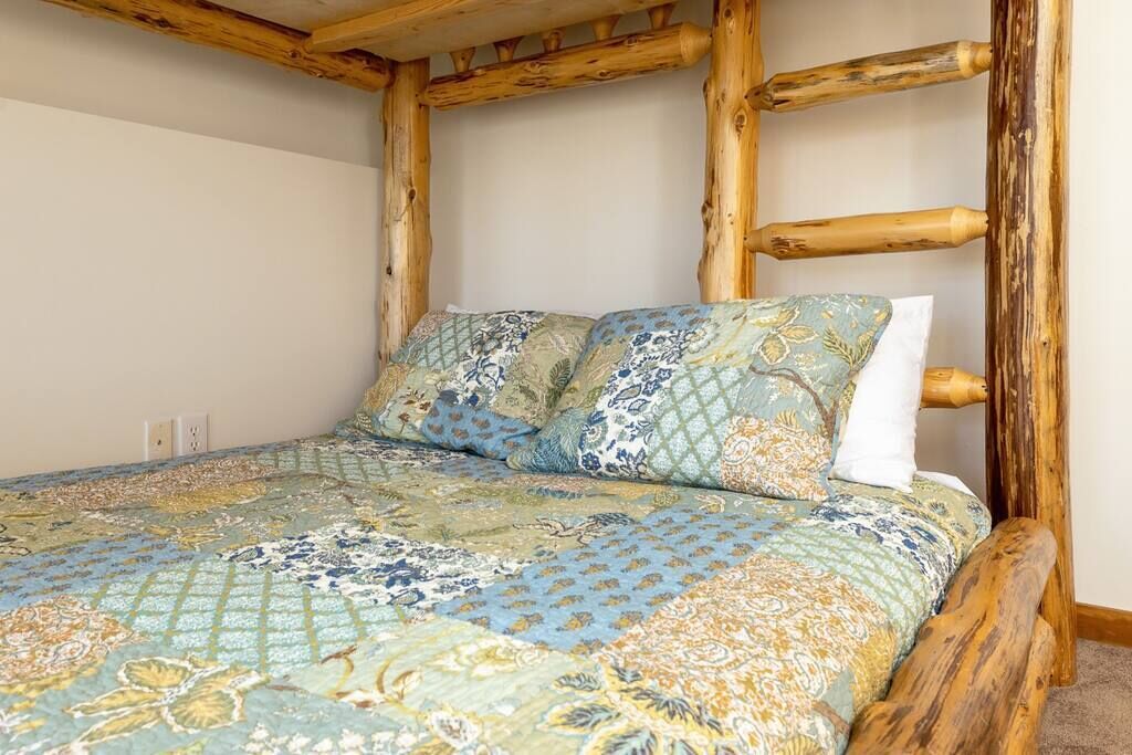 Un lit recouvert d’une couvre-lit à motifs floraux, avec deux oreillers, adossé à une tête de lit en bois, dans une pièce aux murs unis et où une prise électrique est visible.