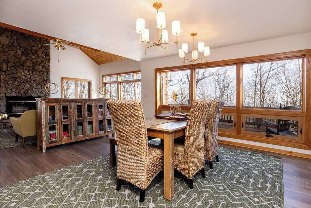 A dining area with a stone fireplace, wicker chairs, a wooden table, and large windows with a view of trees.
