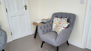 A grey armchair with a floral pillow, a small wooden side table with books, and a white door in a room with beige walls and carpet. - 1 Bed retreat-Sleeps 2-Secure parking-Garden (Ennis)