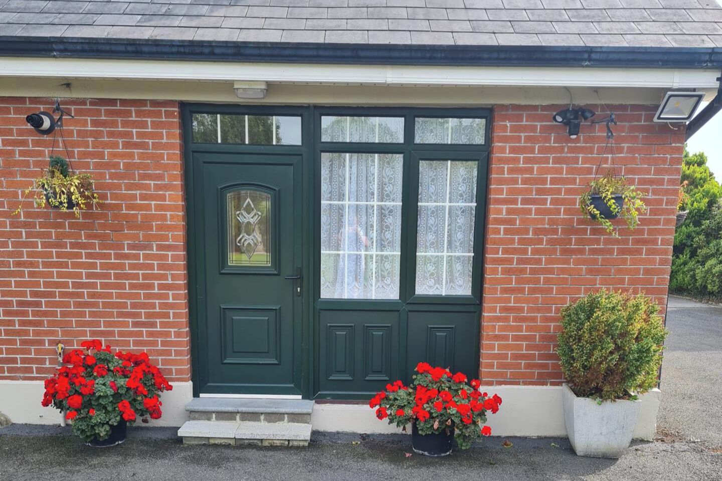 Una casa de ladrillo con una puerta verde, dos macetas con flores rojas y un escalón que lleva a la puerta.