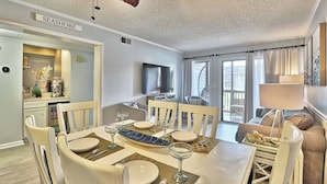 A dining area with a table set for four, a television in the background, and a view of the outdoors through large windows. - Beach & Nightlife 3BR/2.5 BA service dog friendly (North Myrtle Beach)