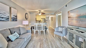 A living room with a gray sofa, a gray armchair, a dining table with chairs, a TV, and a kitchen in the background. - Beach & Nightlife 3BR/2.5 BA service dog friendly (North Myrtle Beach)