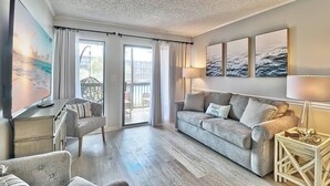 A living room with a grey sofa, a small table with a lamp, a TV, and a painting of a beach scene. - Beach & Nightlife 3BR/2.5 BA service dog friendly (North Myrtle Beach)