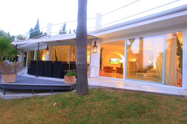 A modern house with a covered outdoor seating area, a large glass door leading to a living room, and a well-maintained lawn. - 2042 Elegant Villa with Garden, heated Pool and Spacious Layout (Marbella)