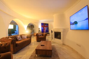 A spacious living room with a fireplace, two sofas, a coffee table, and a large TV screen displaying a scenic view. - 2042 Elegant Villa with Garden, heated Pool and Spacious Layout (Marbella)