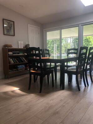 Un coin repas avec une grande table et quatre chaises, une bibliothèque remplie de livres et une maquette de maison, ainsi qu’une porte sur laquelle est accrochée une note.