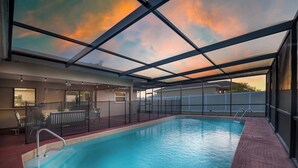 An indoor swimming pool with a glass ceiling, a seating area with chairs, and a view of the sunset.