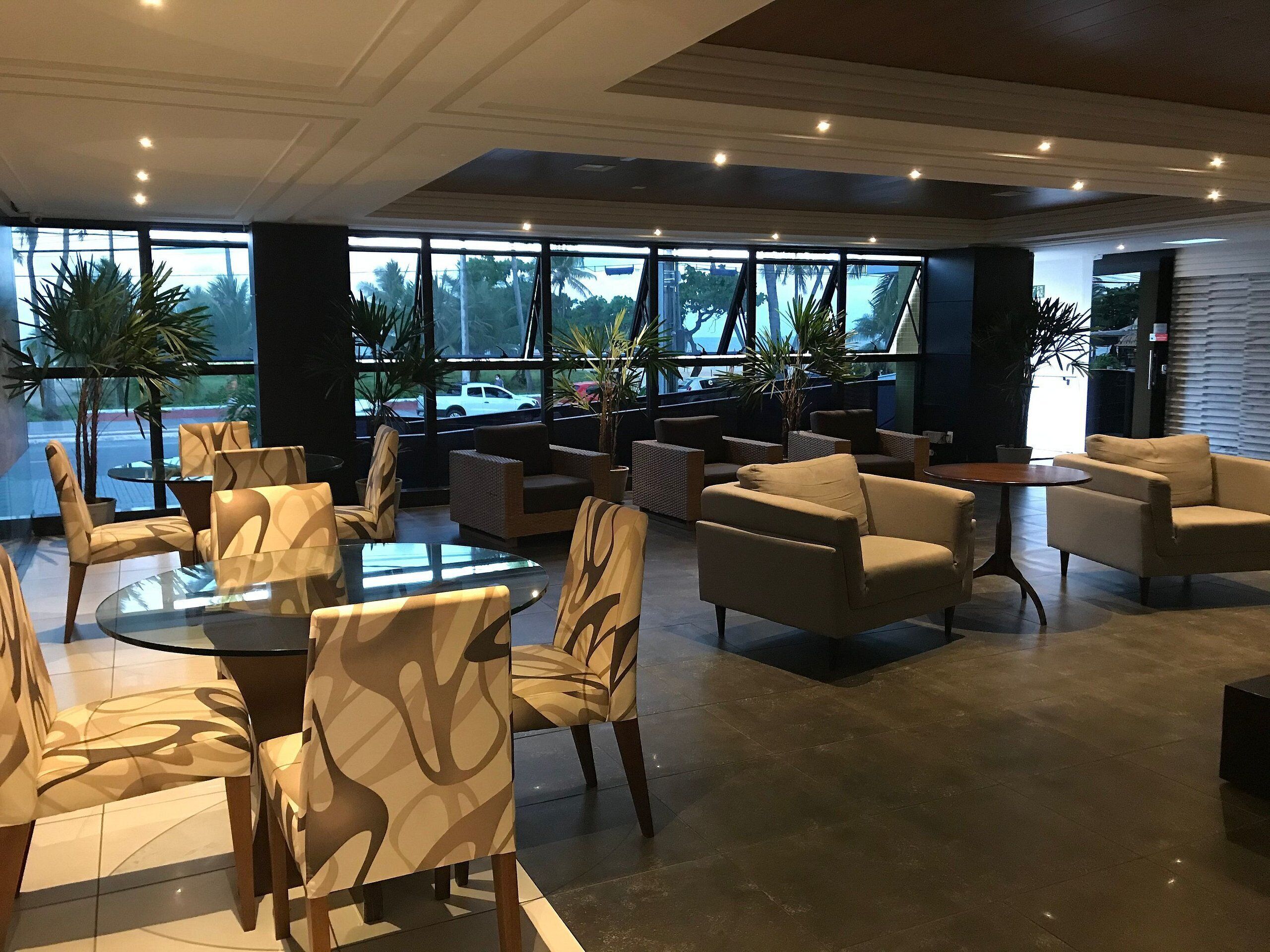 Un área de descanso moderna con una mesa de vidrio, sillas con diseños, sofás y ventanas amplias que dan a vista de palmeras y un estacionamiento.