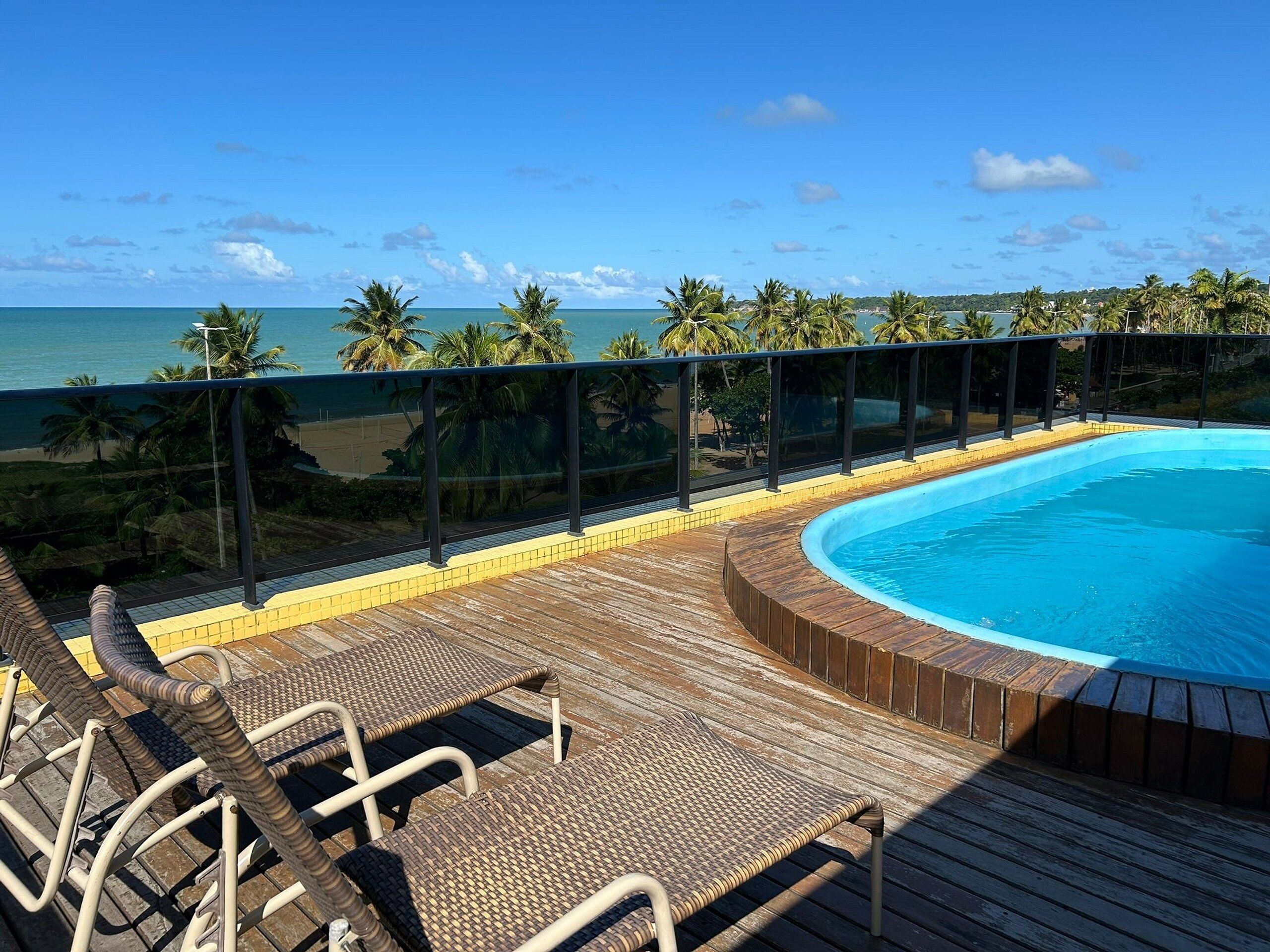 Una piscina con muelle de madera y sillones de descanso, con vista a una playa y al océano.