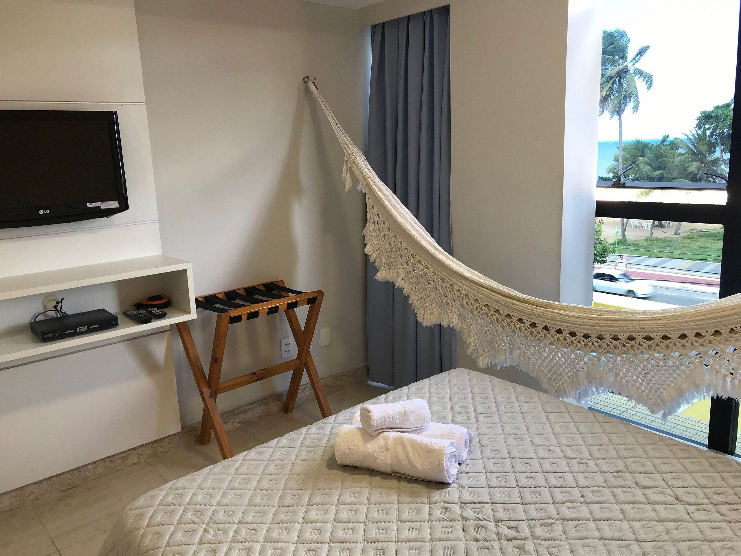 Ein Schlafzimmer mit Bett, Fernseher, kleinem Tisch, Hängematte und Blick ins Freie.