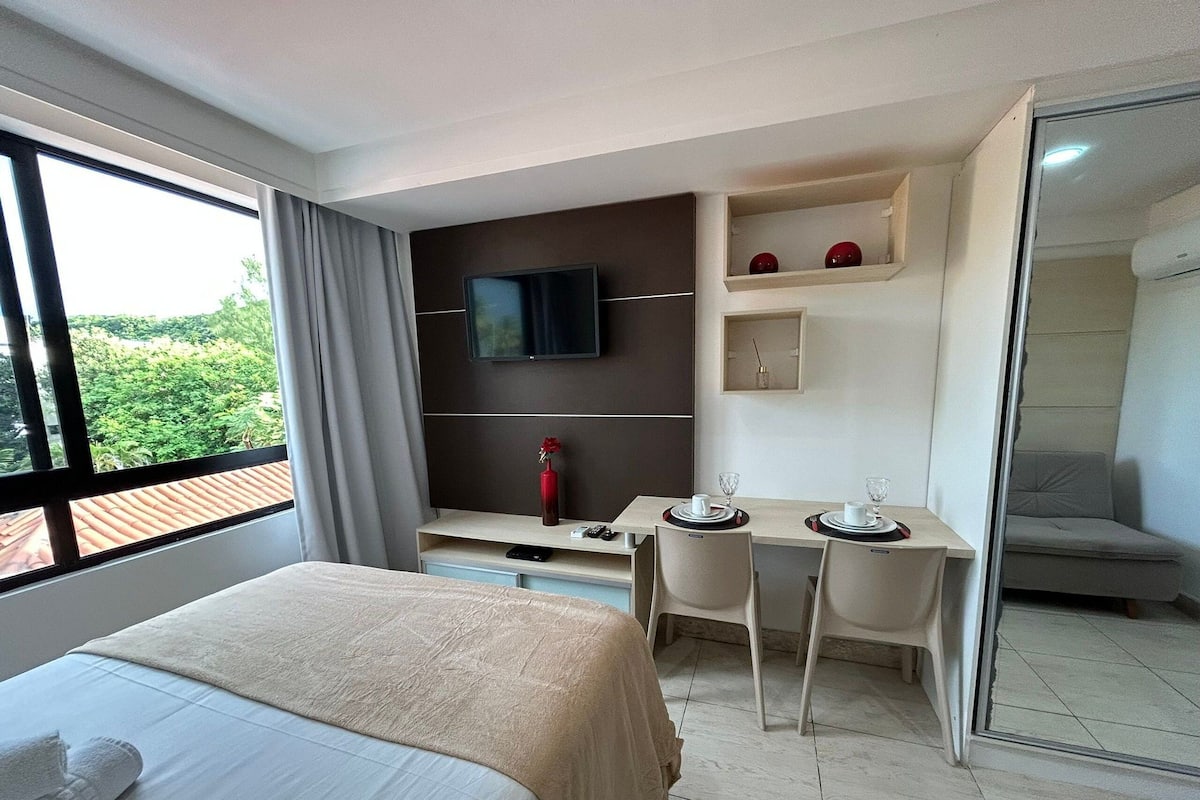 A hotel room with a bed, a TV, a desk with two chairs, and a sliding door leading to a balcony.