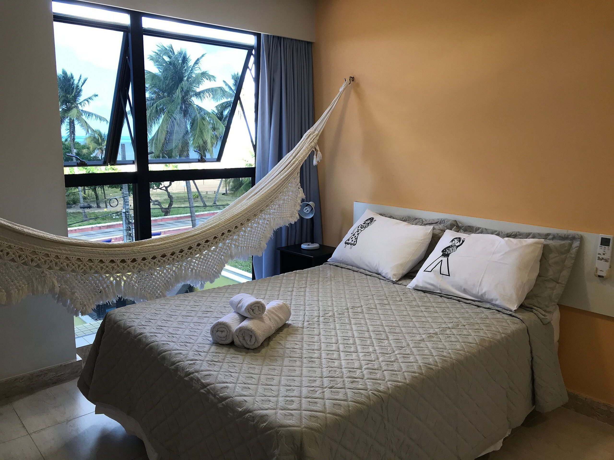 A bed with a quilted cover, two pillows with illustrations, a hammock, and a window with a view of palm trees.