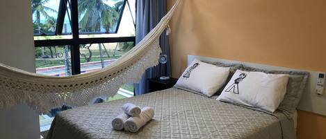 A bed with a quilted cover, two pillows with illustrations, a hammock, and a window with a view of palm trees.