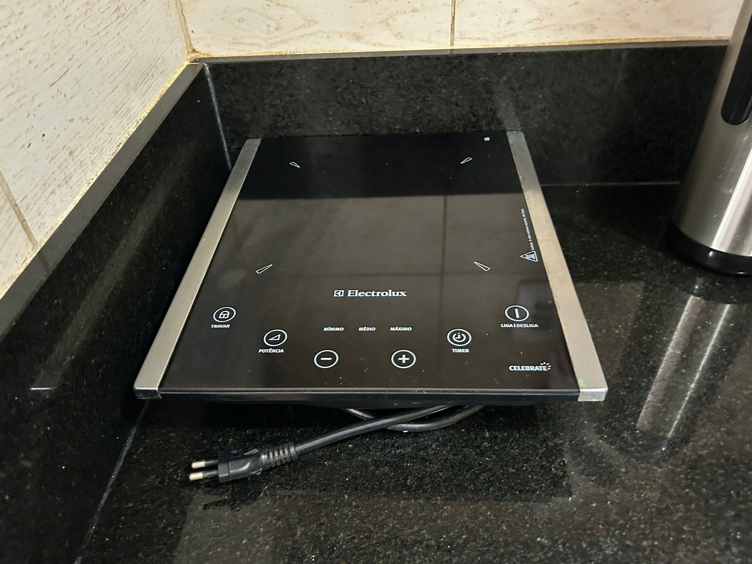 A black induction cooktop with control buttons labeled 'TRAVAR,' 'POTENCIA,' 'MINIMO,' 'MEDIO,' 'MAXIMO,' 'LIGA/DESLIGA,' 'TIMER,' and 'CELEBRATE.'