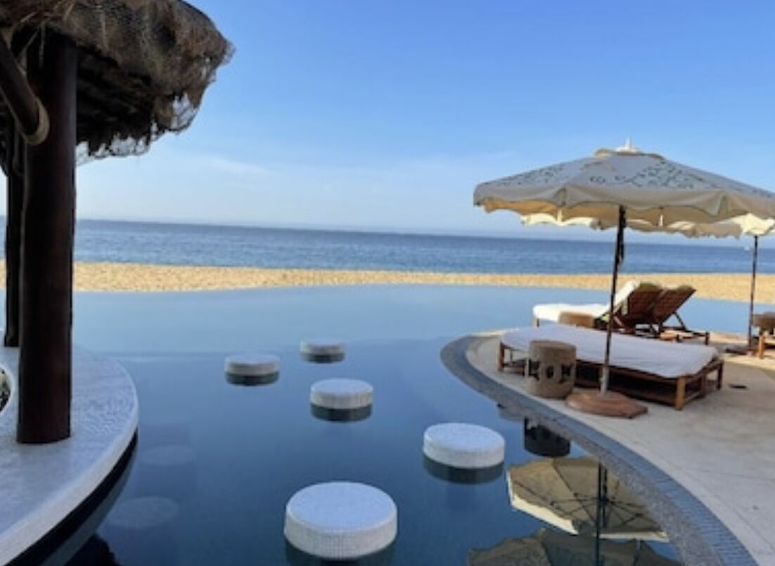 A pool area with circular floating platforms, lounge chairs, and umbrellas, overlooking a beach.