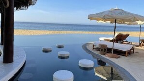 A pool area with circular floating platforms, lounge chairs, and umbrellas, overlooking a beach. - Luxury Villa in 5 Star Resort (Cabo San Lucas)