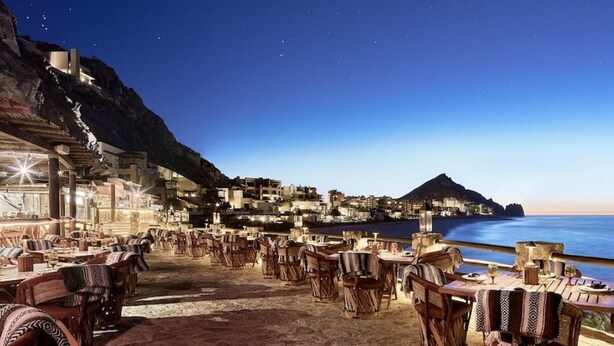 A seaside restaurant with outdoor seating overlooking the ocean, a mountainous backdrop, and a view of a coastal village. - Luxury Villa in 5 Star Resort (Cabo San Lucas)