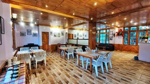 Un restaurante con suelo de madera, sillas y mesas blancas. Hay cuadros en las paredes y un ventanal con vistas a árboles.