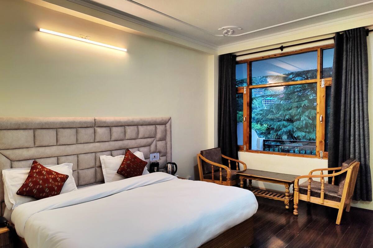 A hotel room with a large bed, a bedside table with a lamp, a chair, and a window with a view of trees.