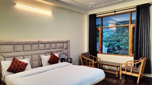 Habitación de hotel con una cama grande, una mesita de noche con lámpara, una silla y ventana con vistas a árboles.