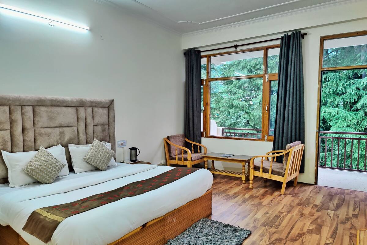 A bedroom with a large bed, a wooden nightstand, a table with chairs, and a window with a view of trees.