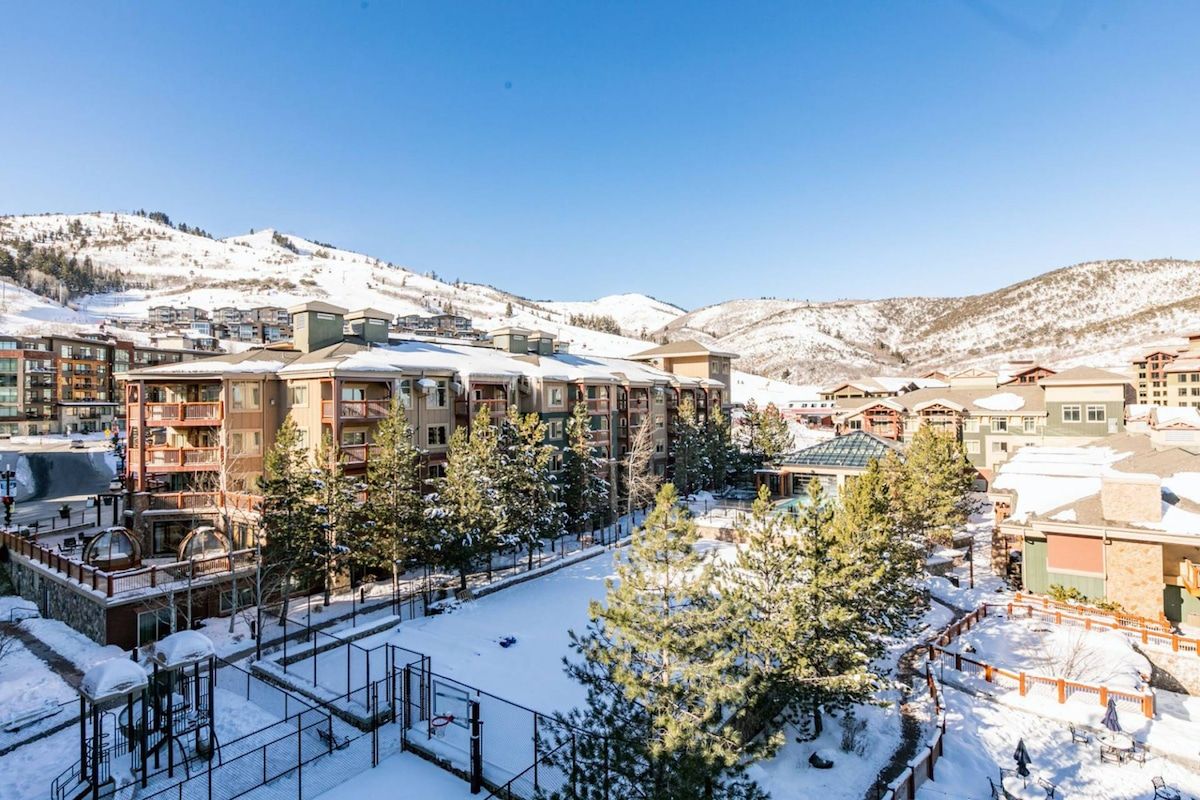 A snow-covered resort with multiple buildings, including a hotel with balconies, surrounded by trees and mountains.
