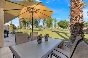 An outdoor patio with a table, chairs, an umbrella, and a view of a well-maintained lawn and palm trees. - Near Coachella | Golf Course, Community Pool & Tennis Courts | Resort Lifestyle (Palm Desert)