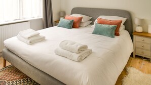 A neatly made bed with white linens and multiple pillows in various colors, a bedside table with a lamp, and a window with curtains.