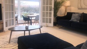 A living room with a dark sofa, a round coffee table, and a TV. There is a door leading to a balcony with a table and chairs outside.