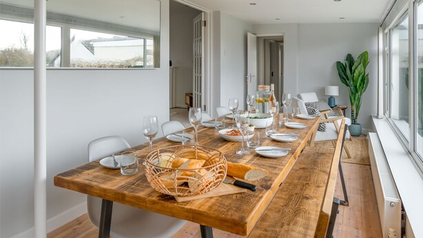 A dining area with a long wooden table set for a meal, white chairs, and a view of the outdoors through large windows.