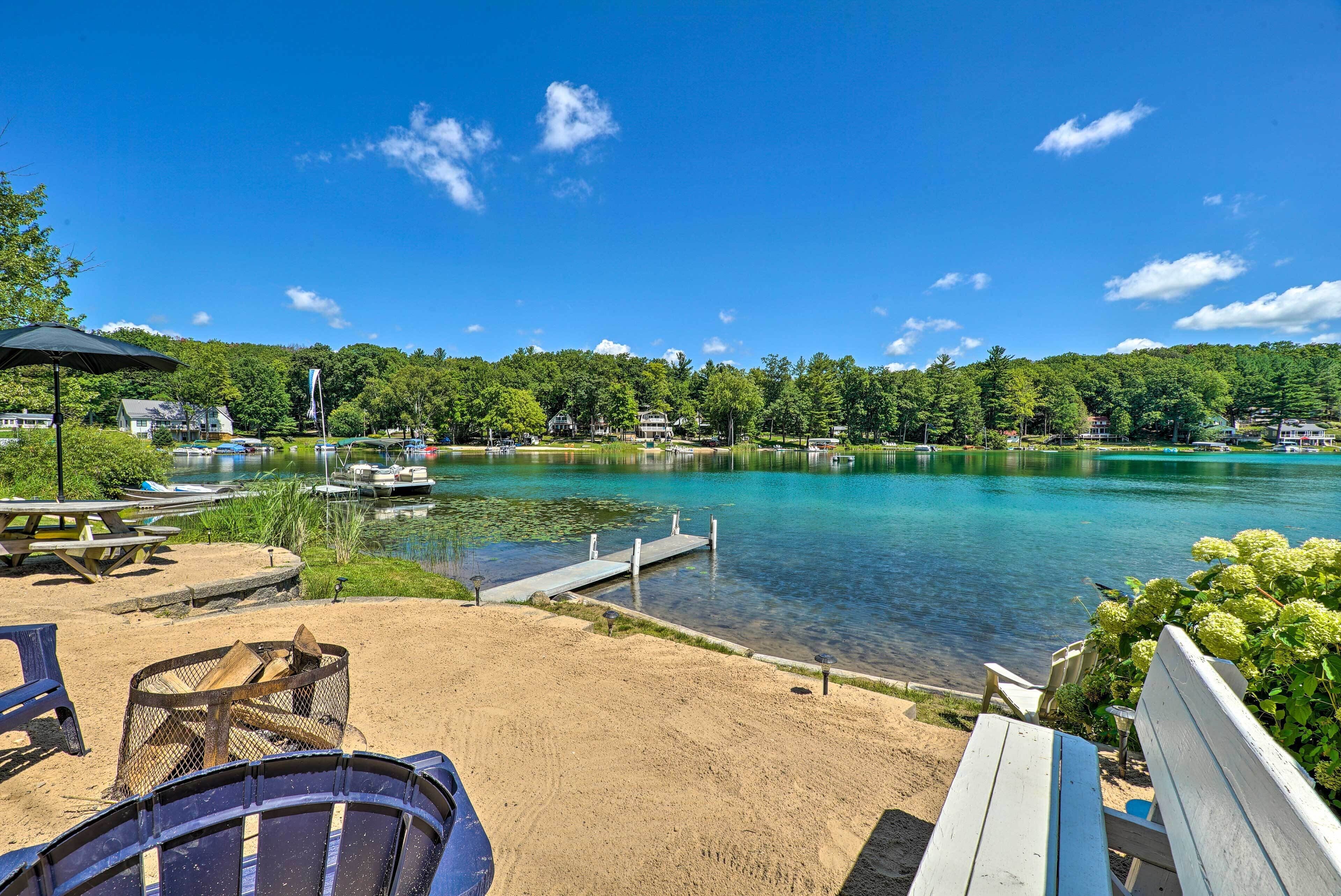 A lakeside view with a dock, picnic tables, and a fire pit.