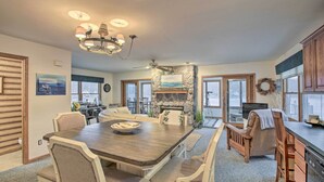 A spacious dining area with a large wooden table and white upholstered chairs, a stone fireplace, and a view of the outdoors through large windows. - Lakefront Newaygo Home - Private Dock, Kayaks (Newaygo)