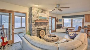 A living room with a stone fireplace, a ceiling fan, a large window, and a view of the outdoors. - Lakefront Newaygo Home - Private Dock, Kayaks (Newaygo)