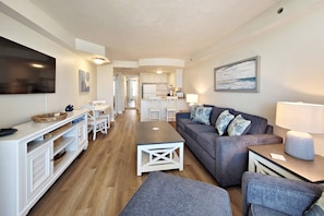 Een moderne woonkamer met een grijze bank, een salontafel en een aan de muur bevestigde televisie. In de achtergrond is een keuken te zien met witte kasten en een eethoek met een tafel en stoelen.