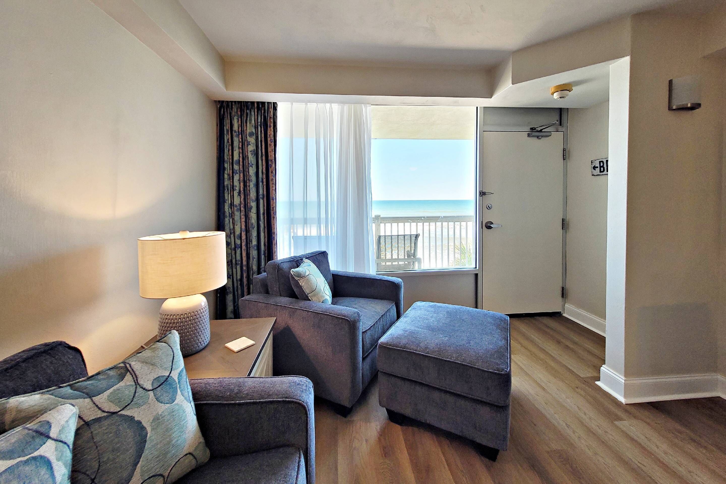 Habitación de hotel con sofá, reposapiés y una mesa con lámpara. Se ve el océano desde la ventana.