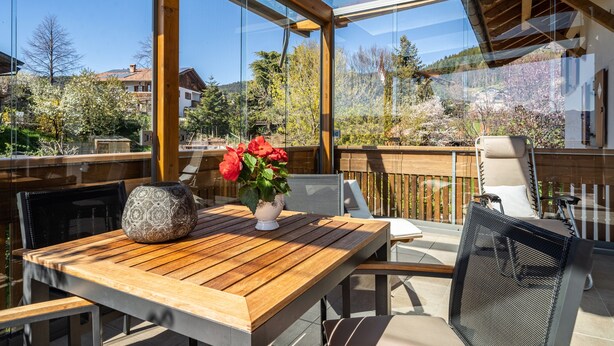 Une table en bois sur laquelle est posé un vase de fleurs rouges, une chaise et une véranda fermée par des vitres, d’où l’on peut admirer des maisons et des arbres.