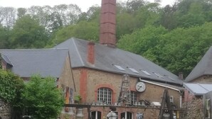 A historic building with a tall chimney emitting smoke, surrounded by greenery and featuring a bell tower. - In the heart of the city Gîte "La cour des trois rois" (The court of the three kings) (Villedieu-les-Poêles-Rouffigny)