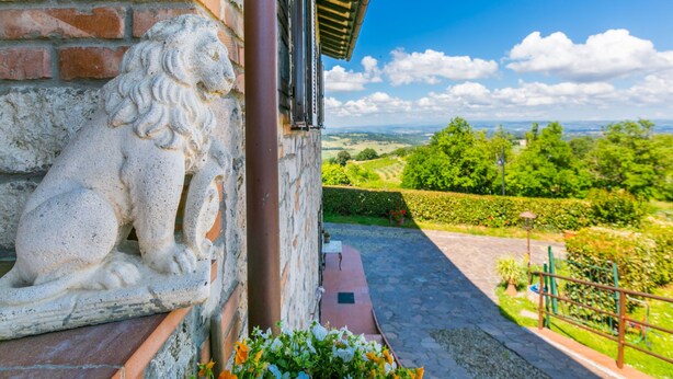 Eine Steintierfigur, ein Löwe, an einer Gebäudefassade, freier Blick auf eine üppige grüne Landschaft und ein gepflasterter Weg, der zu einem Tor führt.
