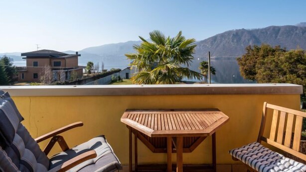 Un balcon avec une table en bois et deux chaises, donnant sur un paysage montagneux et un plan d’eau.