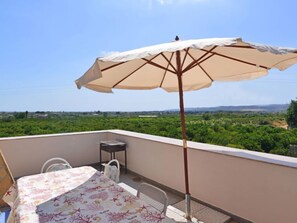 Ein Balkon mit Tisch und Stühlen, einer Tischdecke, einem beige farbenen Sonnenschirm und Blick auf eine grüne Landschaft.