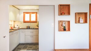 A modern kitchen with white cabinets, a wooden bench, and three wooden display shelves with items. - Vacation home Dorine by Interhome (Nendaz)