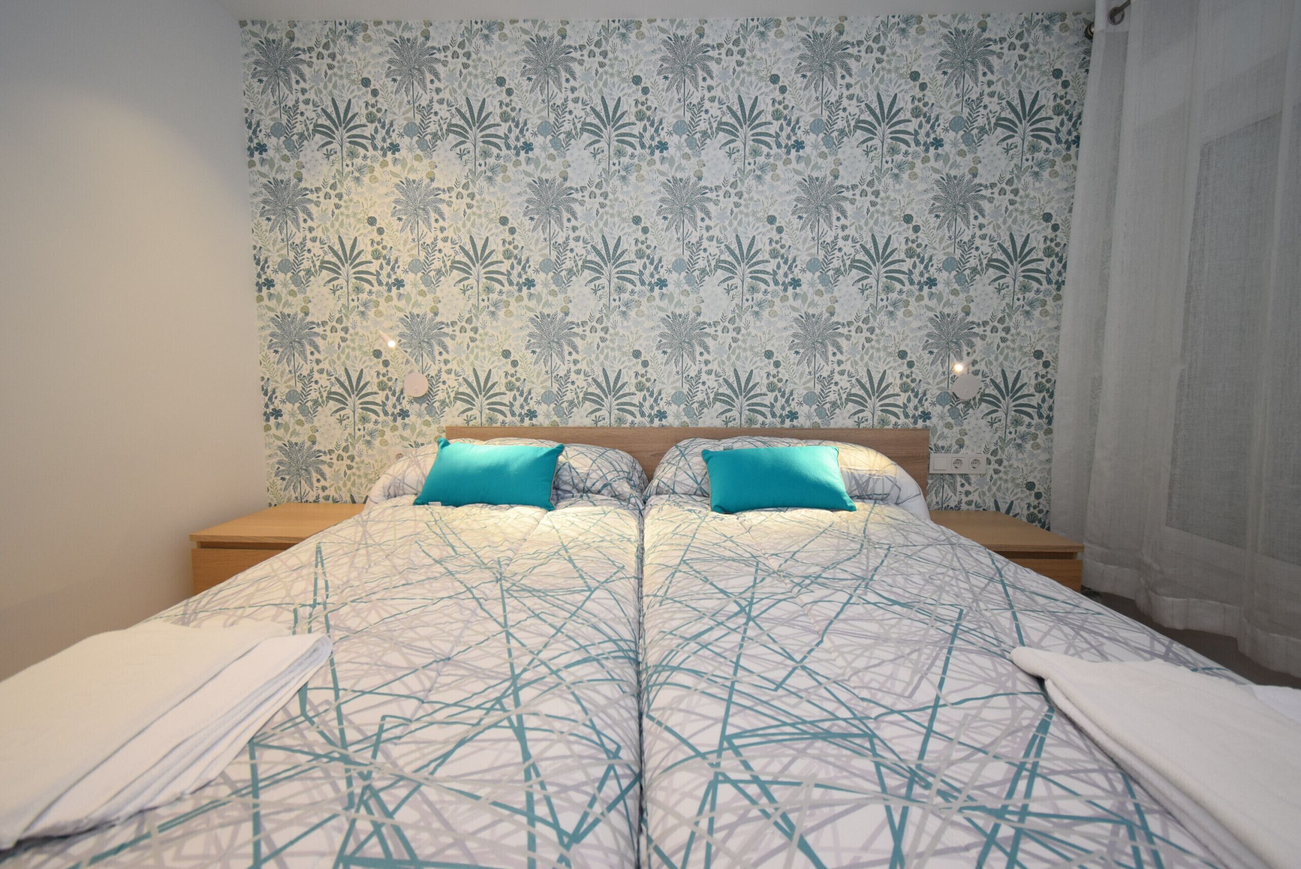Una cama bien hecha con una colcha de estampado geométrico, dos almohadas y una pared con papel tapiz floral.